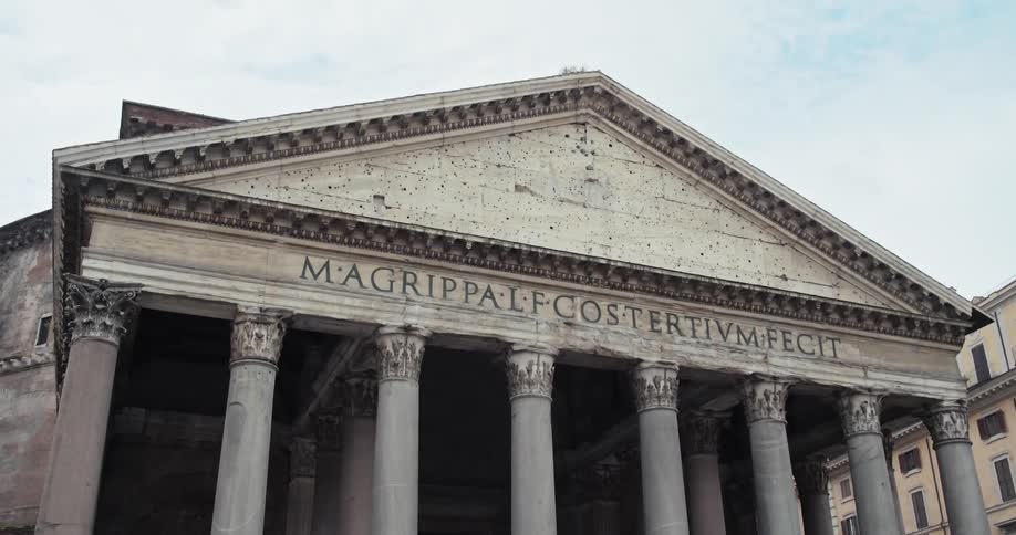 A direct eyelevel shot of the pantheons portico highlighting its massive corinthian columns and the prominent inscription on the triangular pediment all constructed from weathered gray stone