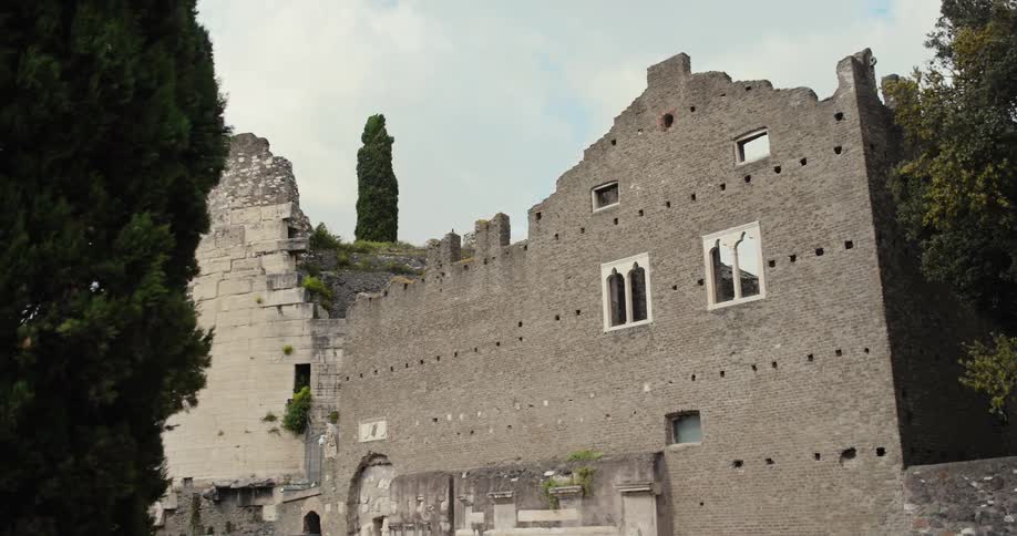 An ancient stone facade of a fortress or ruin with a prominent round tower on the left and tall cypress trees framing the scene against an overcast sky