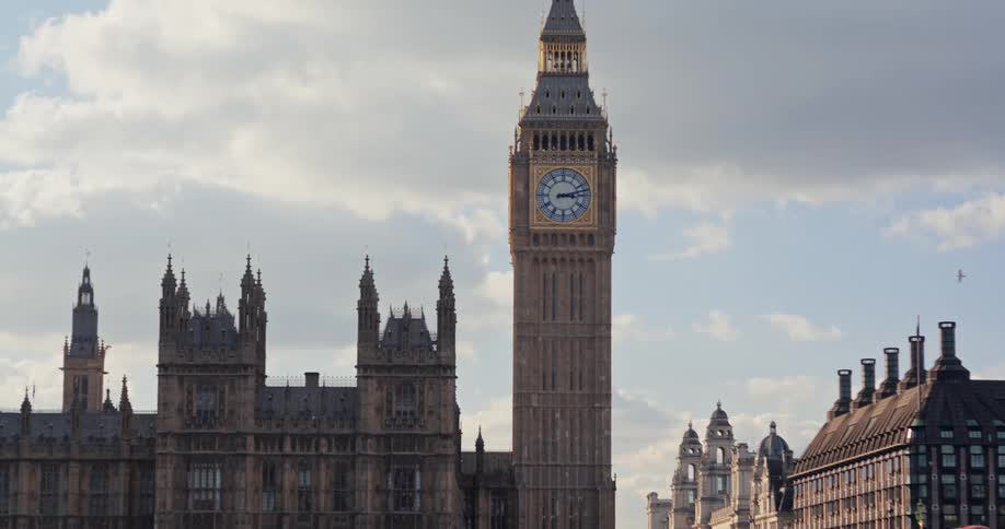 The video shows a view of the palace of westminster and big ben from across the river thames in london the river is visible in the foreground with the stone westminster bridge and its arches crossing it in the background the detailed gothic architectu