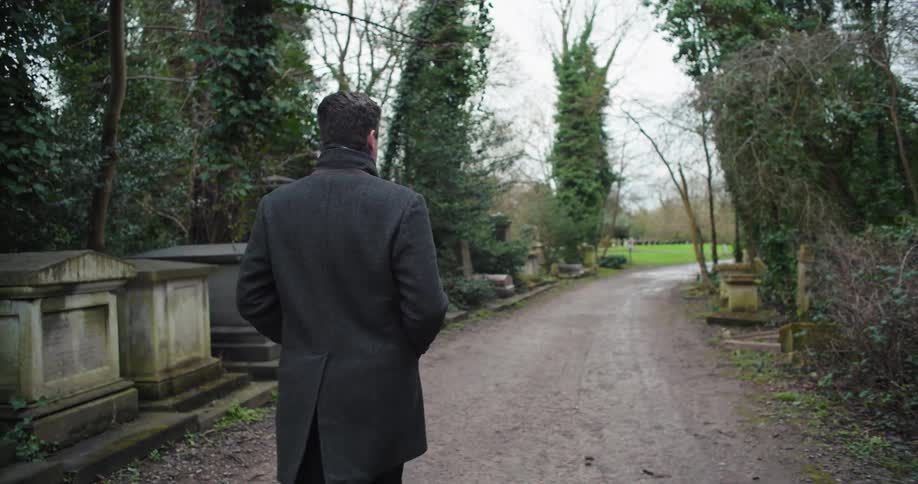 A man walking in a graveyard wearing a jacket surrounded by gravestones and trees