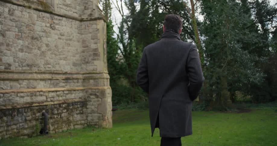A man wearing a jacket coat walking outdoors on a lawn surrounded by trees at midday