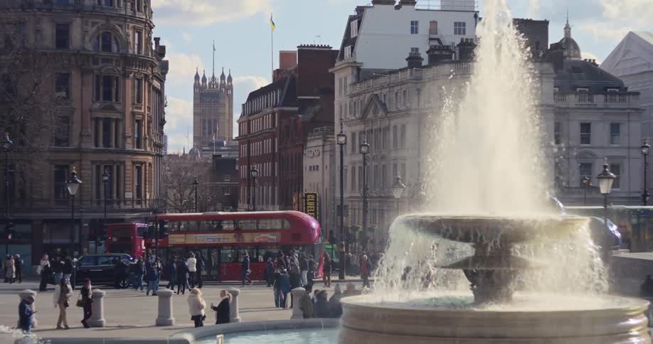 A fountain in the middle of the city sprinkling water as people pass by with a bus driving along the busy road at midday