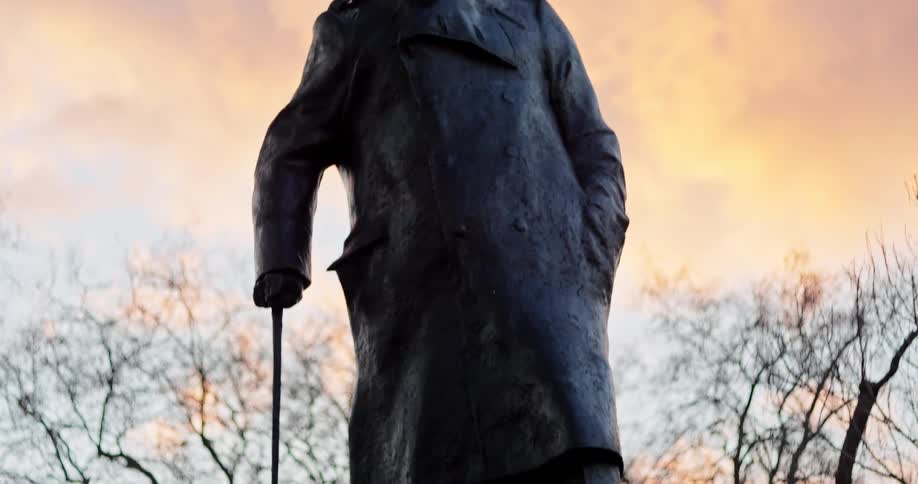Statue of churchill stands in a park near leafless trees under red tinted clouds