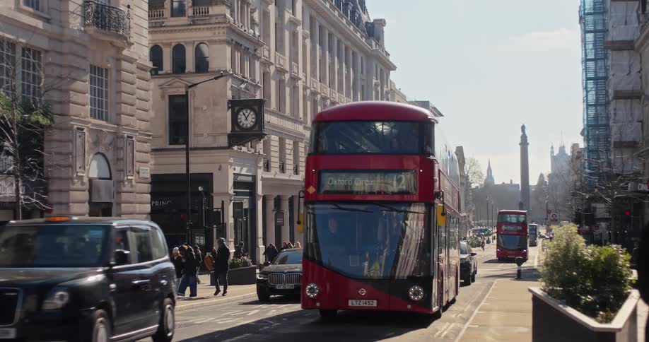 A detailed shot of two red doubledecker buses moving along a sunny city street lined with stately buildings with a classic black taxi in the foreground