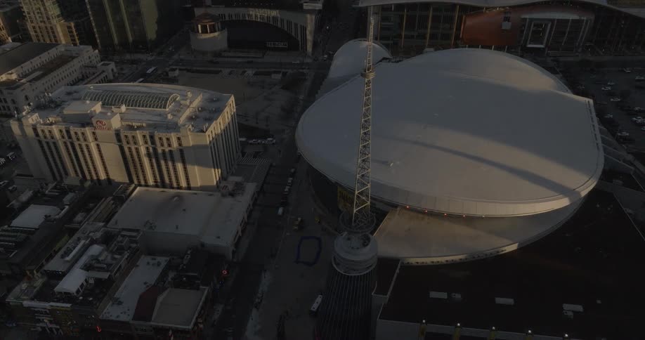 The camera slowly pans over a large white building with a curved roof surrounded by other city buildings and a busy street under a gray overcast sky