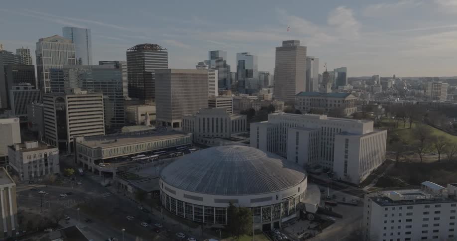 An aerial view of a city shows a large dome shaped building in the foreground with a city skyline and a park visible in the background under a sunny blue sky