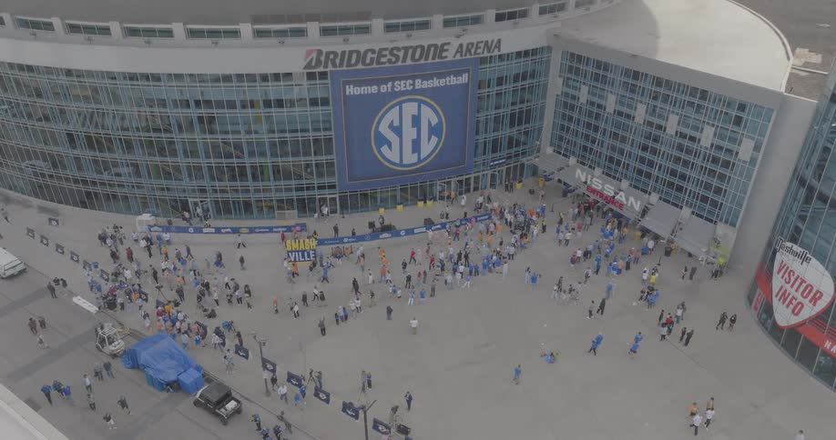 A large crowd gathers outside bridgestone arena in nashville during the day for an sec basketball event with fans in colorful attire filling the plaza