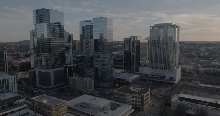 A modern city skyline with many tall glass buildings rises against a hazy sky with a golden light with streets and smaller buildings visible in the foreground