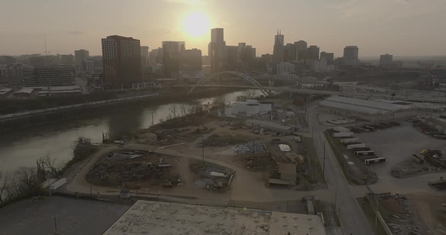 The setting sun creates a bright glare behind a large city skyline which rises above a river and a bridge in the foreground with an industrial area below