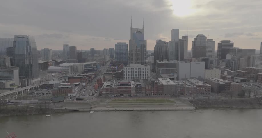 A tall city skyline with a distinctive building in the center rises above a river and a tiered grassy embankment under a cloudy sky