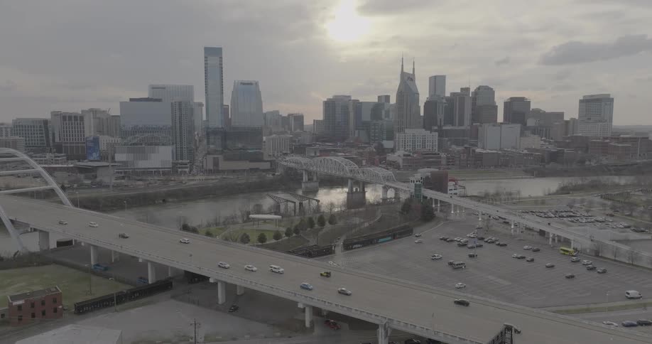 A large city skyline with numerous tall buildings is visible behind a river and two bridges with a large parking lot and busy streets in the foreground under a cloudy sky