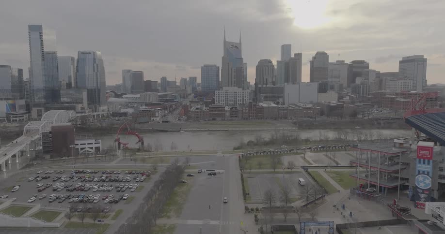 A tall city skyline with a distinctive building in the center rises above a river and a tiered grassy embankment under a cloudy sky