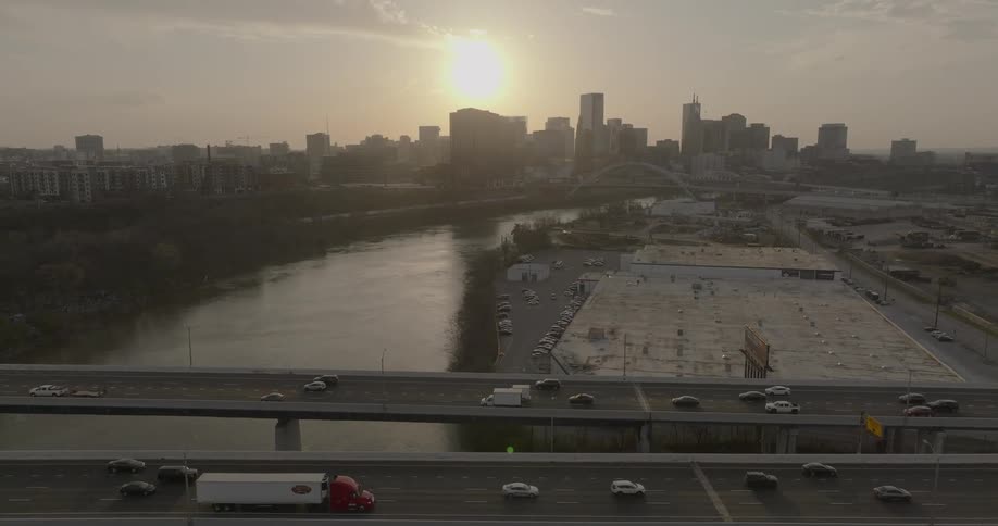 The setting sun creates a bright glare over a city skyline and a river with two parallel bridges and a highway full of cars in the foreground