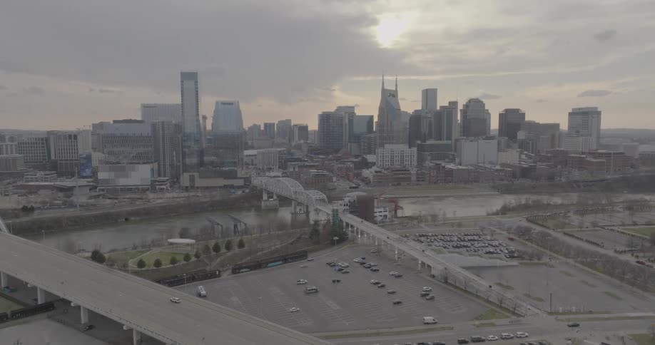 A city skyline of tall buildings is visible behind a river and multiple bridges with a large parking lot and an empty street in the foreground under a cloudy sky