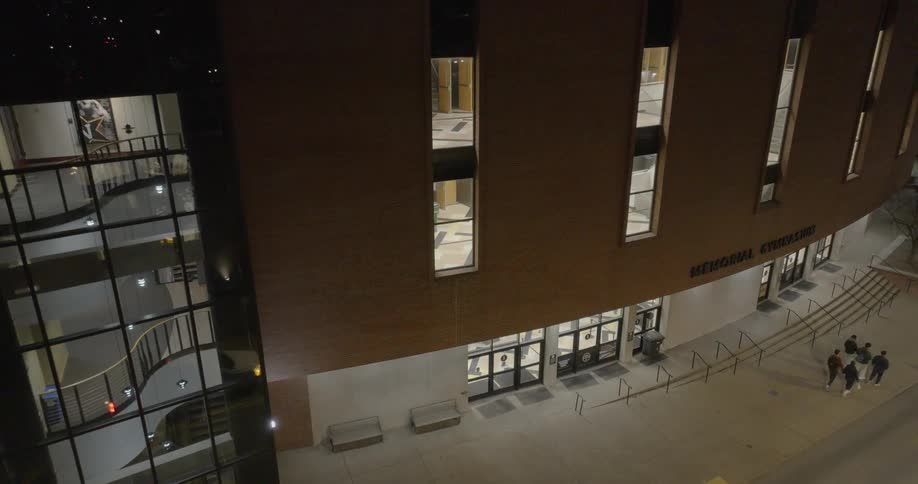 The camera slowly zooms in on a large brown brick building with a curved facade and lit windows as a small group of people walks out of the main entrance at night