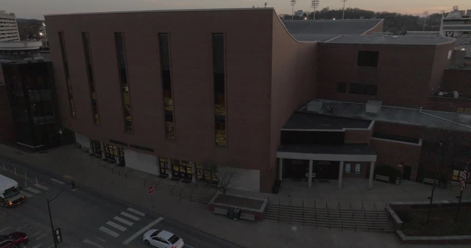 A large brown brick building with a curved facade and tall windows stands on a city street at dusk with a white shuttle bus driving past it