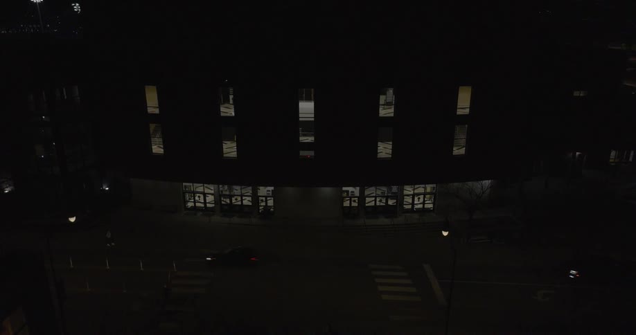Cars going by on the street at night passing a building with lights reflecting out of the windows