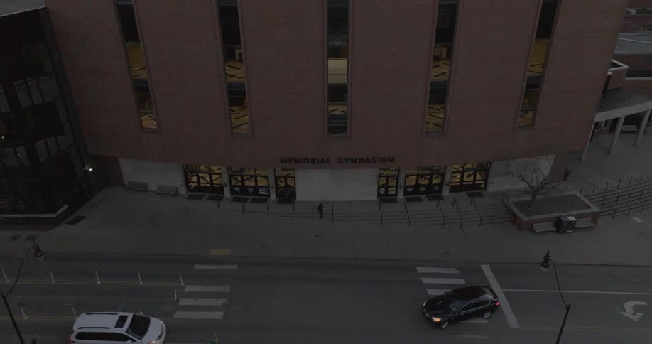 Cars pass by memorial gymnasium as pedestrians walk along the sidewalk in the early evening