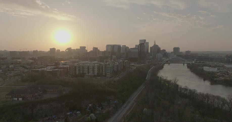 The nashville skyline is silhouetted against a glowing sunset with the cumberland river and john seigenthaler pedestrian bridge visible in the distance