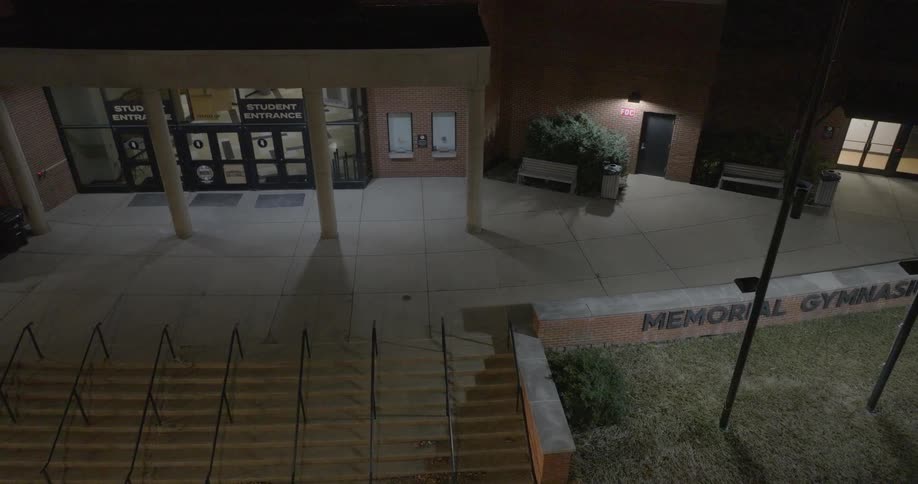 Steps lead past the flagpoles and the wall labeled memorial gymnasium to the student entrance at night