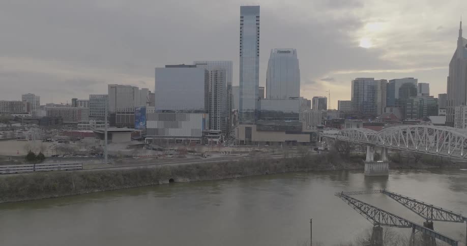 An aerial shot of downtown shows a city skyline and a bridge spanning a river as a train moves along the riverbank on a cloudy day