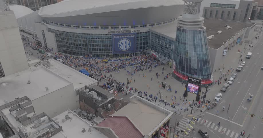 An aerial shot shows a large crowd of people gathered outside a modern arena the bridgestone arena with a large sec banner displayed prominently on its facade surrounded by busy city streets and other downtown buildings