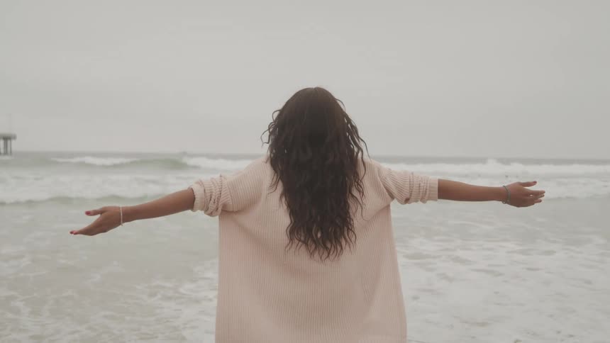 A woman facing the sea at the beach contemplating the waves with her arms open