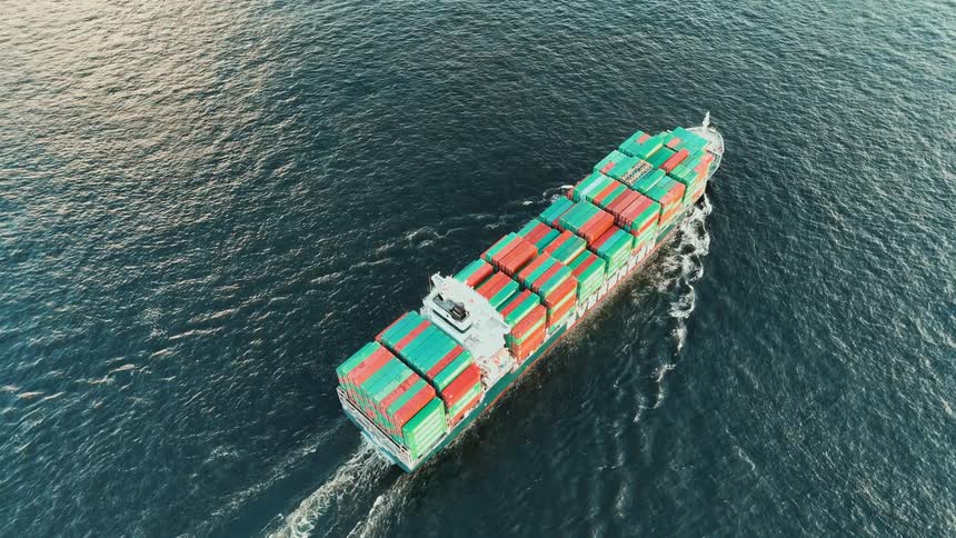 An aerial view shows a large green cargo ship carrying orange and green containers as it moves through the deep blue water leaving a white wake behind it