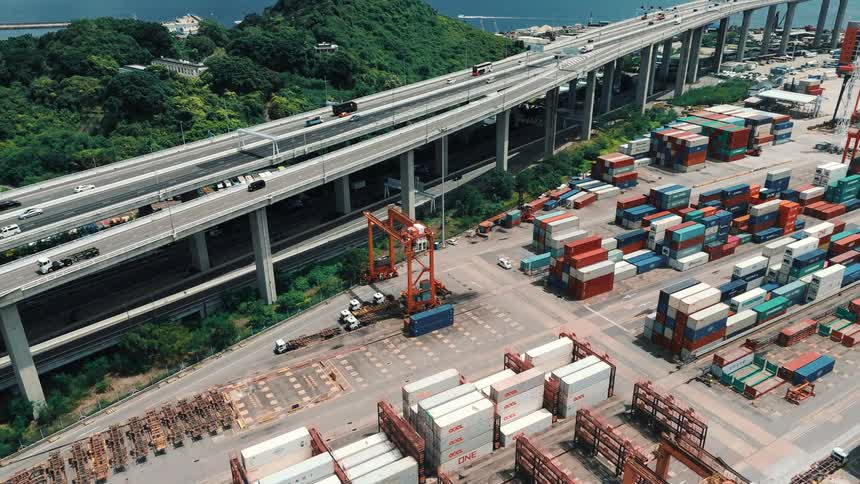 Shipping containers are stacked in a busy port yard beneath an elevated highway curving around a lush green hill