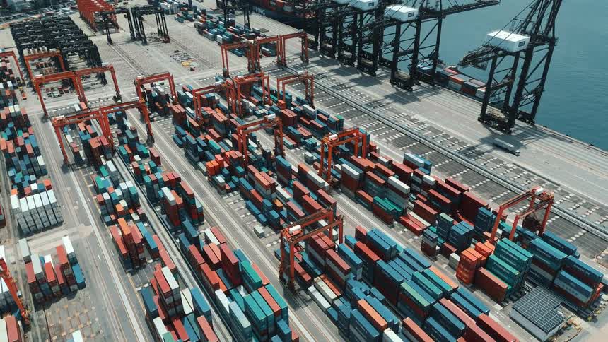 A seaport filled with containers stacked on top of each other at midday with cars and shipping trucks driving by