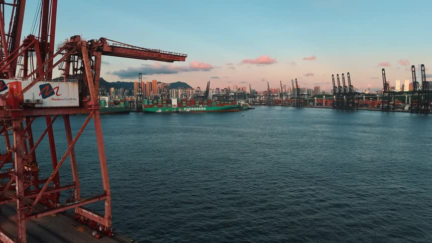 A panoramic shot of a bustling port at twilight framed by two large red cranes with a green cargo ship docked in the foreground and a dense city skyline backed by mountains in the distance