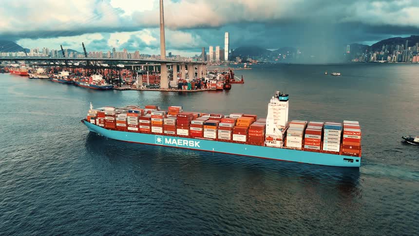 A maersk cargo ship sails through hong kong harbor in daylight with stonecutters bridge city skyscrapers and a looming rainstorm in the distance