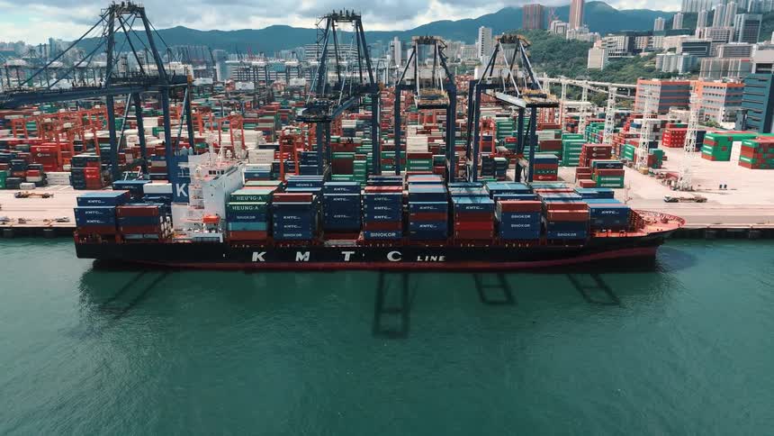 A large cargo ship with many colorful containers on its deck is docked at a port with cranes and stacks of containers behind it all next to a city with green hills
