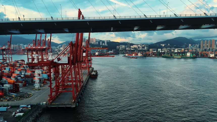 Port terminal with red cranes and stacks of containers under a bridge with a city view in the background