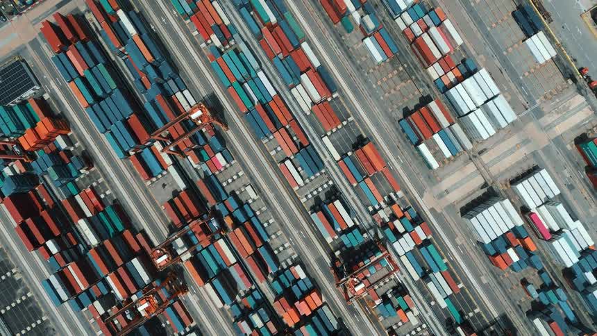 An aerial topdown view of a massive shipping terminal showcasing a dense colorful grid of stacked shipping containers with large gantry cranes moving between them