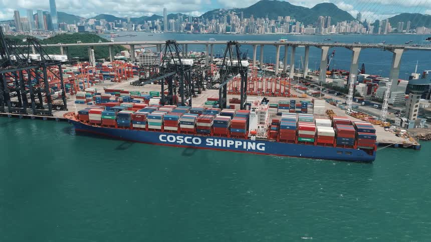 A massive cosco shipping container vessel is docked at a bustling port with cranes unloading cargo against the backdrop of a modern coastal city and distant mountains