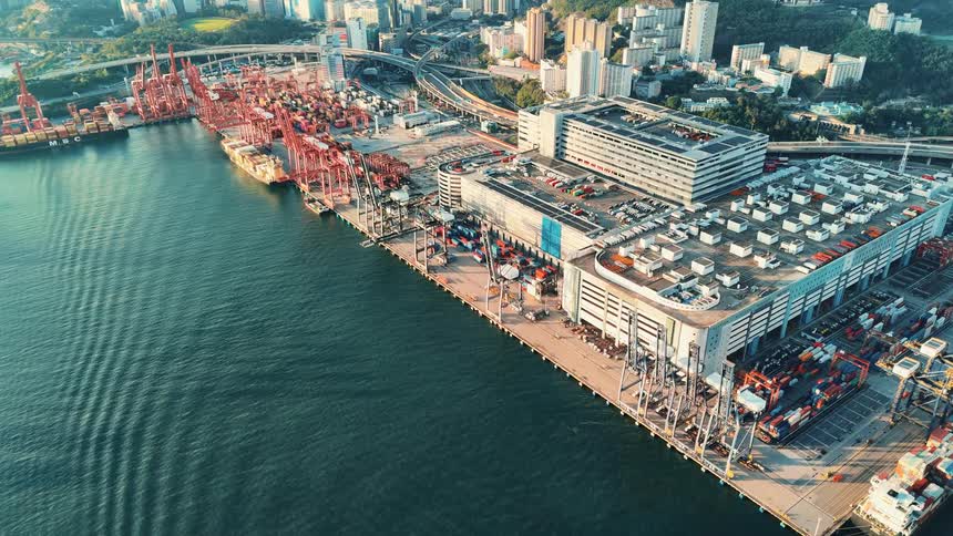An aerial shot captures a sprawling industrial port bustling with red cranes and large cargo ships docked along the waterfront with a dense city and network of roads visible in the background