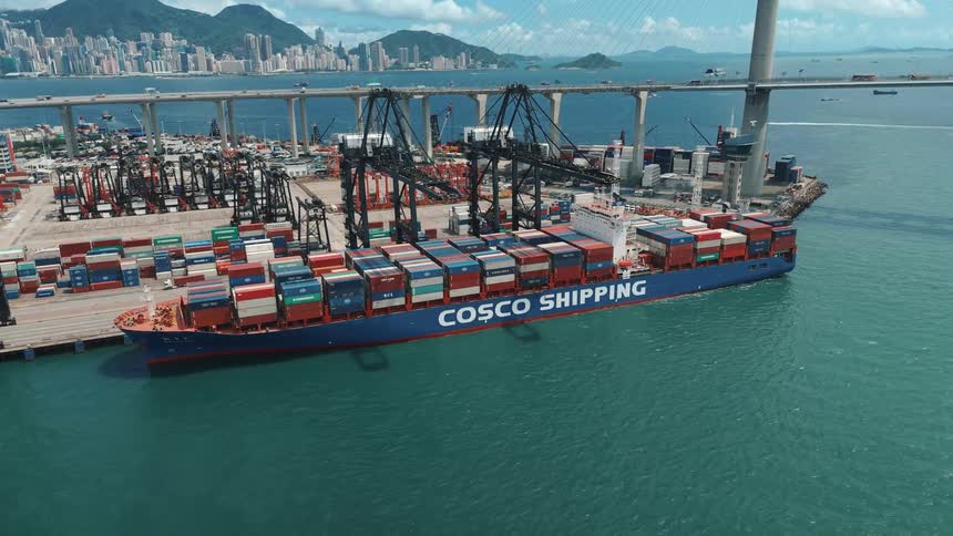 An aerial view of a large container ship docked at a busy port with numerous cranes and stacked containers set against a backdrop of a massive bridge a sprawling city skyline and mountains under a bright cloudy sky