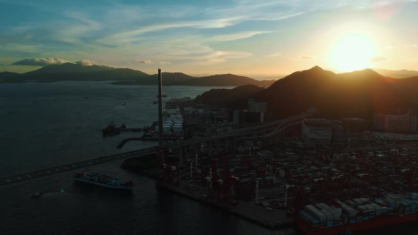 A busy port with many cranes and shipping containers sits at the base of a mountain as the sun sets behind it with a suspension bridge and a ship on the water