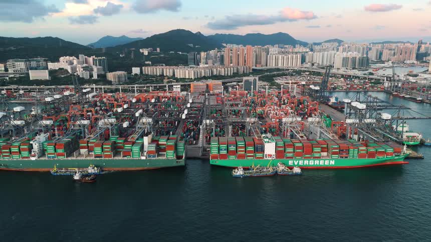 An aerial shot captures the complex largescale industrial activity of a container port with massive ships being worked on against a backdrop of a dense urban skyline and rolling hills