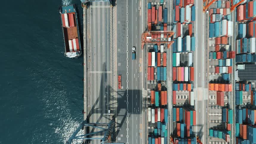 An aerial view of a bustling shipping port during the day shows a cargo ship being unloaded by cranes beside rows of colorful stacked containers
