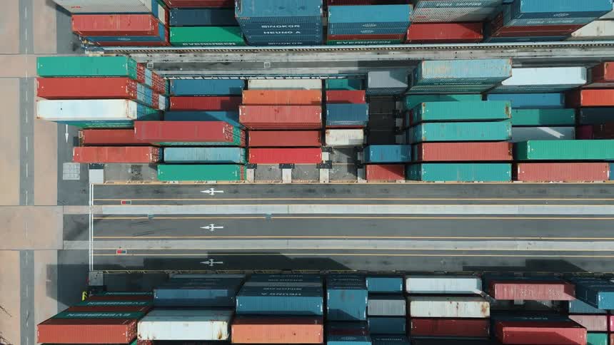 Shipping containers are stacked in a busy port yard beneath an elevated highway curving around a lush green hill