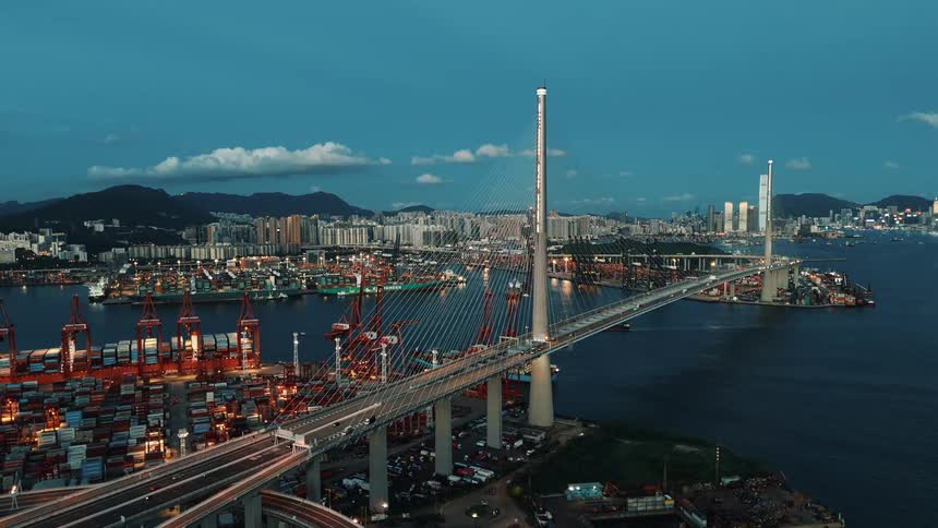 An aerial shot at twilight captures a massive cablestayed bridge spanning a harbor with a sprawling brightly lit container port in the foreground and a dense city skyline backed by hills in the distance