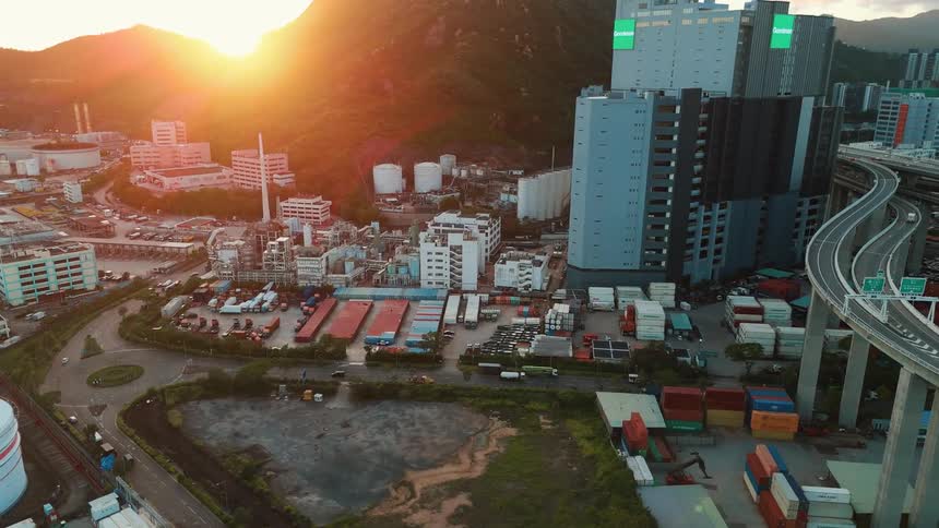 A busy industrial area with many buildings storage tanks and shipping containers is in the foreground of a city with a large curving highway and a mountain in the background under a setting sun