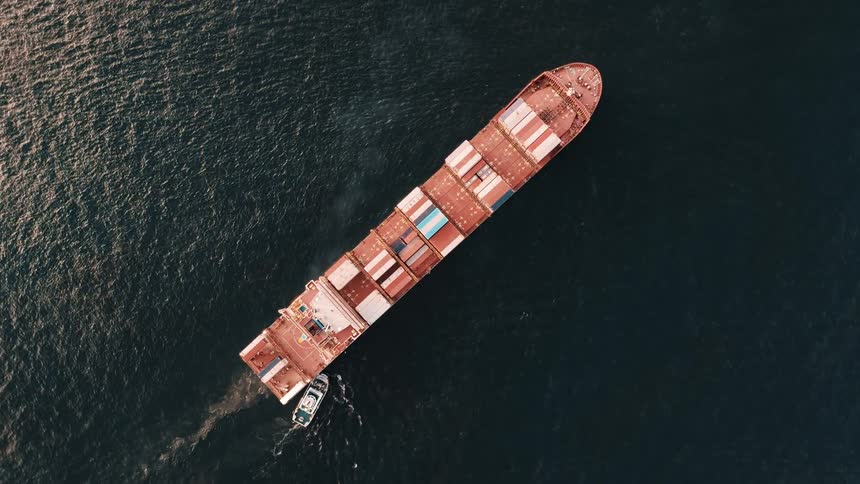An aerial view shows a large red cargo ship carrying colorful containers with a smaller white boat beside it moving through the deep blue water