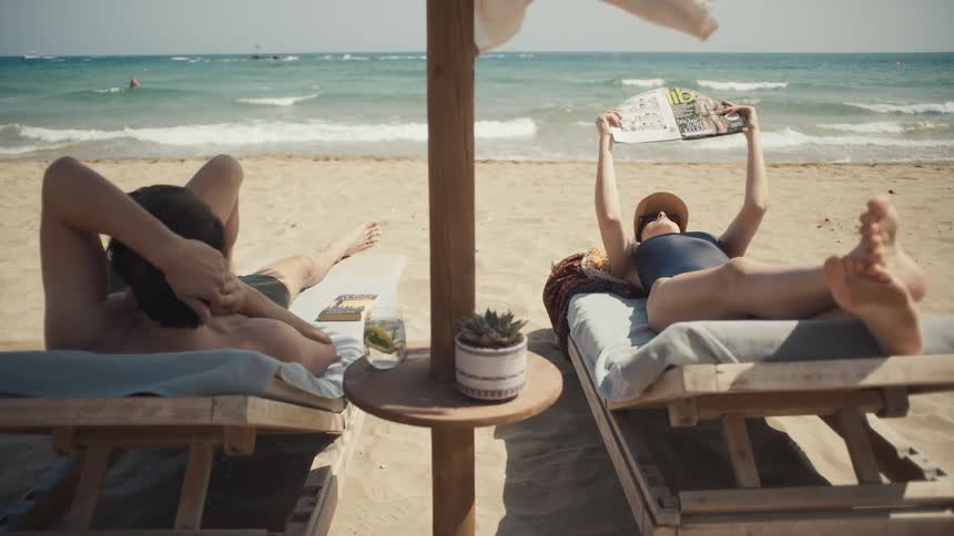 Two people lying on beach loungers by the sea under a wooden sunshade