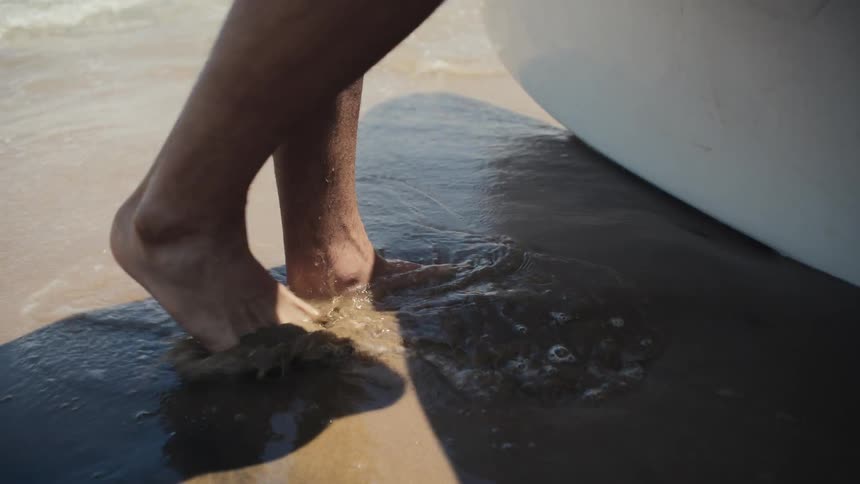 A persons bare feet stand on a beach as waves wash over them on the sandy shore