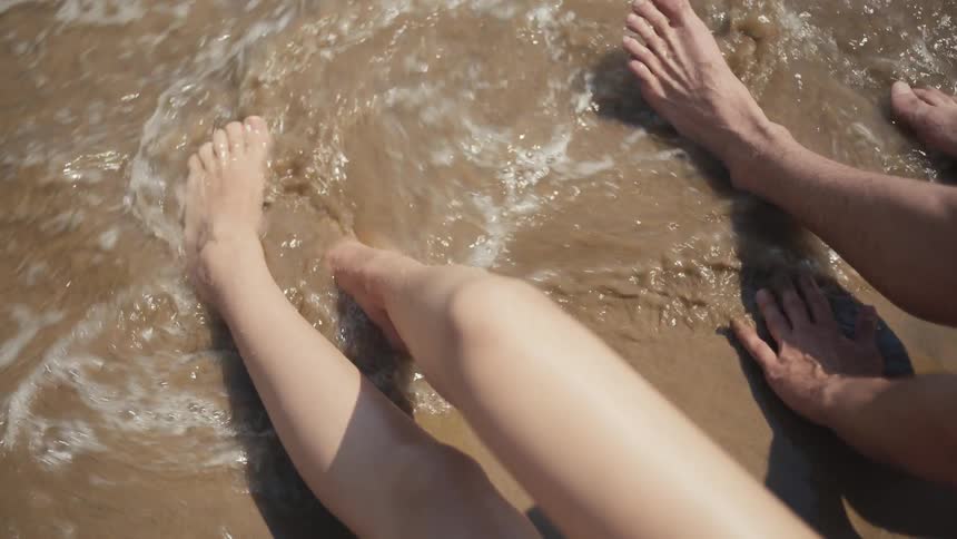 Two people sit closely together on wet sand at the beach their bare legs stretched out and partially overlapping in the sunlight