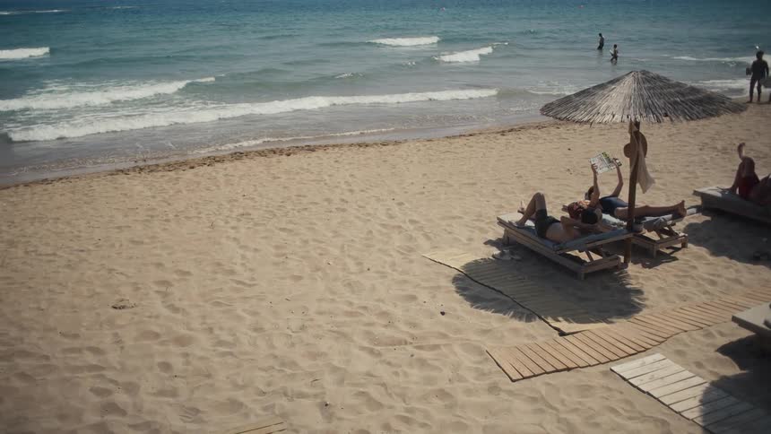 A sandy beach with lounge chairs and a thatched umbrella sits next to the ocean where people are swimming with others lying on the chairs under a bright blue sky