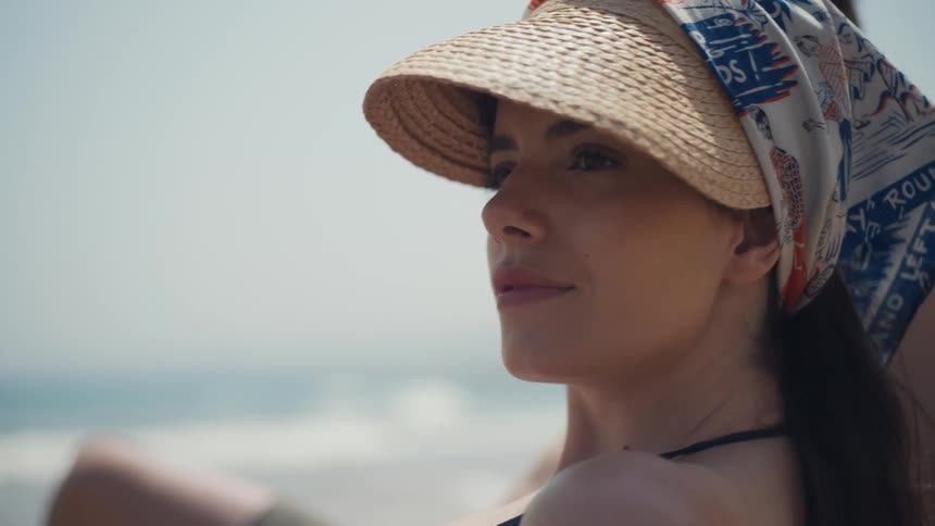 Woman wearing a straw hat and patterned scarf sitting by the sea